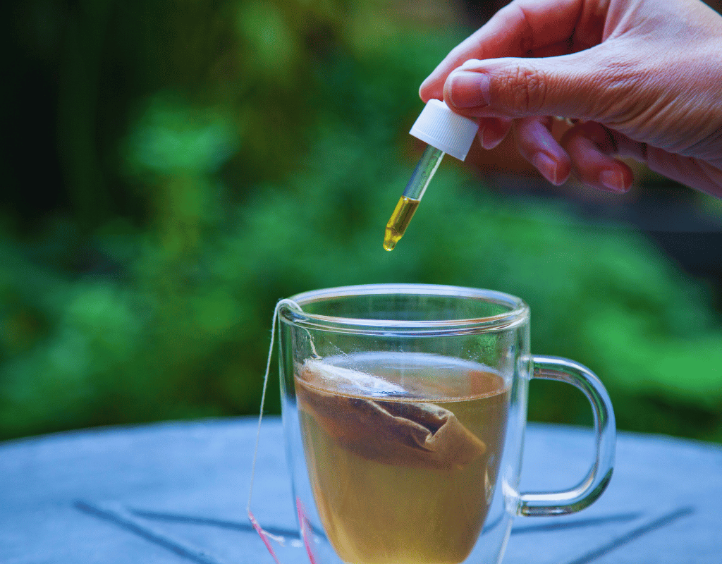Hand hält Pipette mit gelbem Tropfen über Tasse mit hellem Tee, grüner Hintergrund
